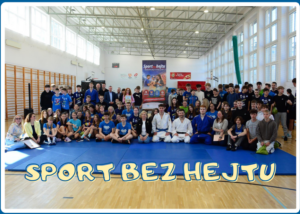 Duża grupa dzieci, młodzieży i dorosłych pozuje do wspólnego zdjęcia w jasnej hali sportowej. Uczestnicy siedzą i stoją na niebieskiej macie, część z nich ubrana jest w stroje sportowe, w tym judogi. W tle widoczne są kosze do koszykówki, drabinki gimnastyczne oraz baner promujący aktywność fizyczną. Na dole zdjęcia znajduje się napis „SPORT BEZ HEJTU”.