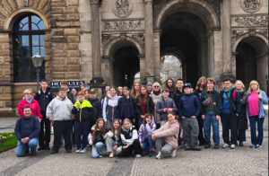 Grupa około 30 osób (głównie dzieci i nastolatków z kilkoma dorosłymi), sfotografowana podczas zwiedzania Drezna, stoi i kuca na brukowanym placu przed monumentalnym, kamiennym budynkiem z ozdobnymi łukami i rzeźbieniami.