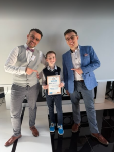 Dwóch uśmiechniętych dorosłych mężczyzn stoi po bokach chłopca i wskazuje na niego, podczas gdy on trzyma w rękach dyplom. Chłopiec ubrany jest w szkolny strój (koszula i kamizelka), a mężczyźni mają eleganckie stroje – jeden w kamizelce i muszce, drugi w marynarce. Cała trójka stoi w jasnym wnętrzu na czarno-białej podłodze, co sugeruje uroczyste wręczenie nagrody lub wyróżnienia.